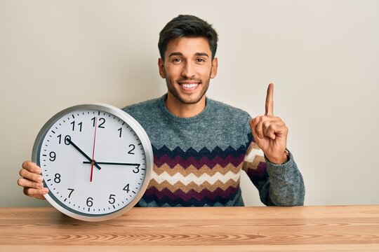 Young handsome man holding big clock smiling with an idea or question pointing finger with happy face, number one