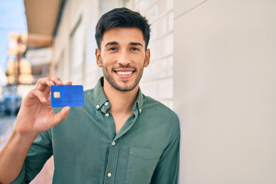 Young latin man smiling happy holding credit card leaning on the wall at the city.