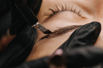 Naklejka premium eyebrow microblading. A master in black gloves holds a blending needle over the brow of the model. Macro photography