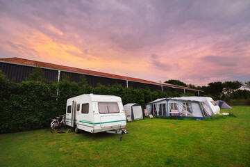 White caravan trailer with a bicycle in a camping site. Spring landscape. Pink sunset clouds....