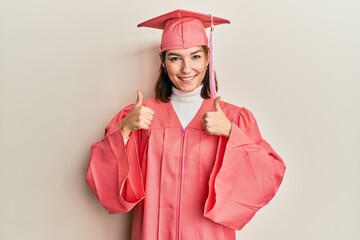 Young caucasian woman wearing graduation cap and ceremony robe success sign doing positive gesture with hand, thumbs up smiling and happy. cheerful expression and winner gesture.