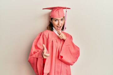 Young caucasian woman wearing graduation cap and ceremony robe pointing fingers to camera with...