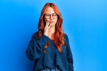 Young beautiful redhead woman wearing casual sweater and glasses over blue background touching mouth with hand with painful expression because of toothache or dental illness on teeth. dentist
