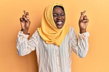Beautiful african young woman wearing traditional islamic hijab scarf gesturing finger crossed smiling with hope and eyes closed. luck and superstitious concept.