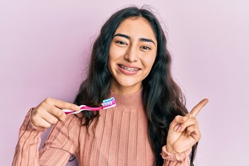 Hispanic teenager girl with dental braces holding mouthwash and toothbrush for fresh breath smiling...