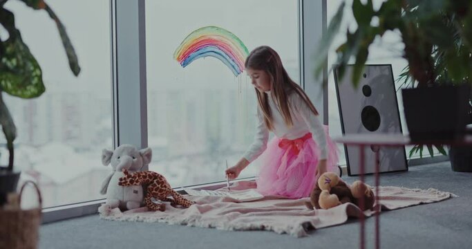 Kids Staying Home. Artistic Little Girl Wearing Fairy Pink Dress Drawing Rainbow Picture on Window and Playing with Toys. Young Generation. Hobbys.