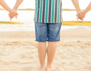 young man in shorts and striped shirt holding hands with his friends in front of the beach. friendship, travel and leisure concept