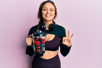 Young hispanic girl wearing sportswear holding food processor mixer machine with fruits doing ok...