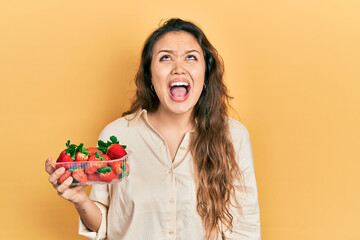 Young hispanic girl holding strawberries angry and mad screaming frustrated and furious, shouting with anger. rage and aggressive concept.
