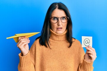 Middle age brunette woman holding email symbol and paper plane in shock face, looking skeptical and...