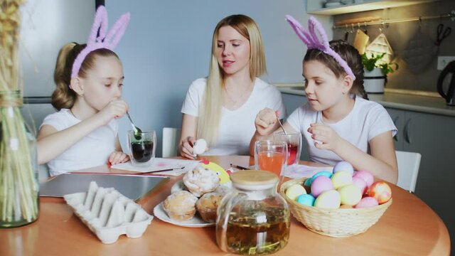 A mother and her daughters painting Easter eggs. Happy family preparing for Easter. Cute little girls wearing bunny ears on Easter day.