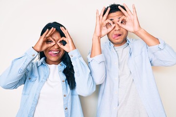 Beautiful latin young couple wearing casual clothes doing ok gesture like binoculars sticking tongue out, eyes looking through fingers. crazy expression.