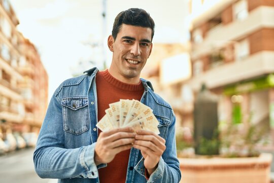 Young hispanic man smiling happy holding denmark krone banknotes at the city.