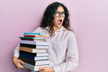 Young brunette woman with curly hair holding a pile of books angry and mad screaming frustrated and furious, shouting with anger. rage and aggressive concept.