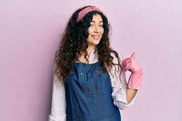 Young brunette woman with curly hair wearing cleaner apron and gloves smiling with happy face looking and pointing to the side with thumb up.