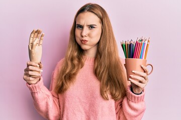 Young blonde woman holding small wooden manikin hand and pencils puffing cheeks with funny face....