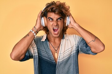 Young hispanic man listening to music using headphones angry and mad screaming frustrated and...