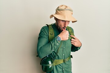 Handsome man with beard wearing explorer hat and backpack feeling unwell and coughing as symptom...