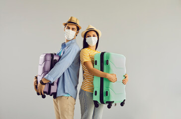 Vacation in pandemic condition, new travel rules. Happy family couple wearing protective respiratory facial mask holding luggage bag looking at camera standing over studio copy space background