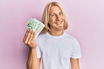 Caucasian young man with long hair holding 20 thai baht banknotes looking positive and happy standing and smiling with a confident smile showing teeth