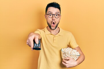 Young hispanic man eating popcorn using tv control afraid and shocked with surprise and amazed...