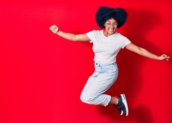 Young beautiful african american woman wearing casual clothes and glasses smiling happy. Jumping with smile on face celebrating with fists up over isolated red background