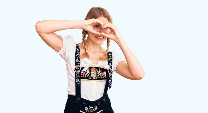 Young beautiful blonde woman wearing oktoberfest dress doing heart shape with hand and fingers smiling looking through sign