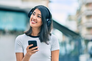 Young hispanic woman smiling happy using smartphone and headphones at the city.