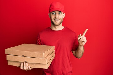 Young caucasian man holding delivery pizza box smiling happy pointing with hand and finger to the side