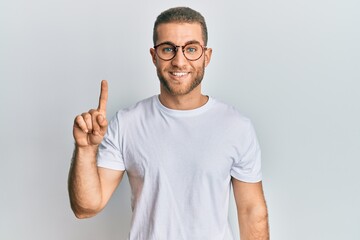 Fototapeta premium Young caucasian man wearing casual clothes and glasses showing and pointing up with finger number one while smiling confident and happy.