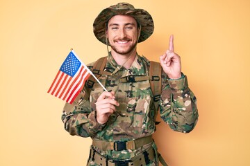 Young caucasian man wearing camouflage army uniform holding usa flag surprised with an idea or question pointing finger with happy face, number one