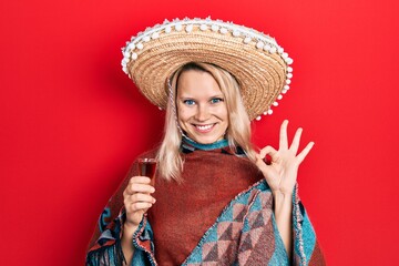 Beautiful caucasian blonde woman wearing festive mexican poncho drinking tequila shot doing ok sign with fingers, smiling friendly gesturing excellent symbol