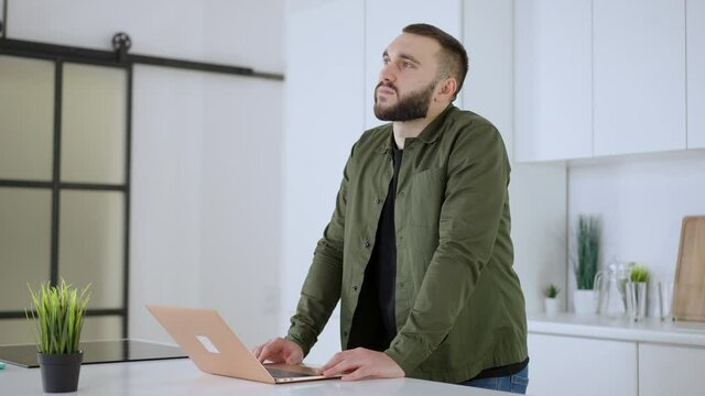Portrait of thoughtful concentrated Caucasian man standing in kitchen with laptop. Bearded young inspired guy thinking of ideas at home indoors. New normal on pandemic lockdown.