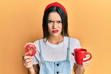 Young hispanic girl eating doughnut and drinking coffee skeptic and nervous, frowning upset because...