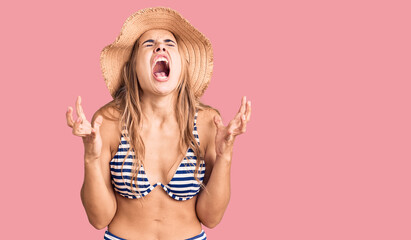Young beautiful blonde woman wearing bikini and hat crazy and mad shouting and yelling with...