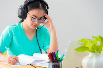 Asian woman student teenage girl with glasses headphones sitting looking serious reading a book worry using laptop computer on table learning online study. Education from a class of university at home