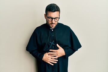 Young hispanic man wearing priest uniform standing over white background with hand on stomach...