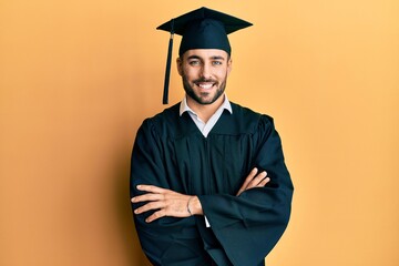 Young hispanic man wearing graduation cap and ceremony robe happy face smiling with crossed arms...