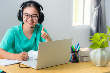 Portrait young Asian female student with headphones glasses smile looking and thumb up a teenage girl is happy to university internet distance study online class works on a laptop computer from home