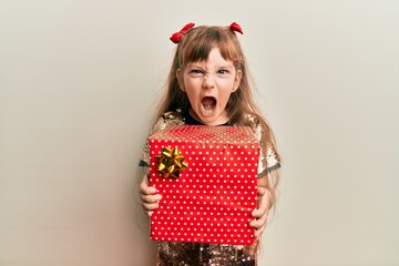 Little caucasian girl kid holding red gift angry and mad screaming frustrated and furious, shouting...