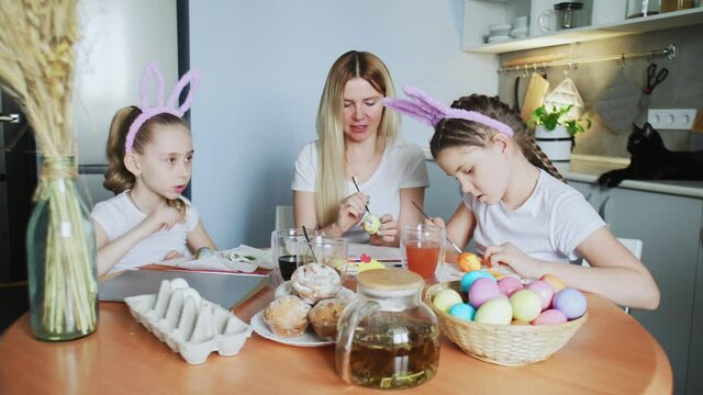 Young mother and her daughters painting Easter eggs. Happy family preparing for Easter. Cute little girls wearing bunny ears on Easter day.