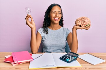 Young african american girl holding brain and light bulb studying for school winking looking at the...