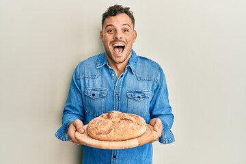 Young hispanic man holding bread celebrating crazy and amazed for success with open eyes screaming excited.