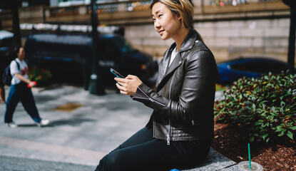 Contemplative female thoughtful looking away while using smartphone technology during touristic vacations, trendy dressed Asian travel blogger with cellphone thinking at urban setting in city