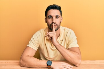 Young hispanic man wearing casual clothes sitting on the table asking to be quiet with finger on...