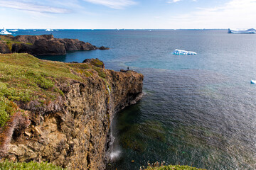 One man standing on rocky cliffs and one waterfall next to blue ocean with floating icebergs and coastline with green moss and grass, Qeqertarsuaq, Greenland, Denmark © vladimir