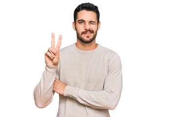 Young hispanic man wearing casual clothes smiling with happy face winking at the camera doing victory sign. number two.