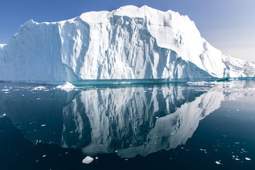 Blueish white iceberg with mirroring reflection in a flat blue cold arctic sea with small floating...
