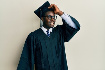 Handsome black man wearing graduation cap and ceremony robe smiling confident touching hair with...