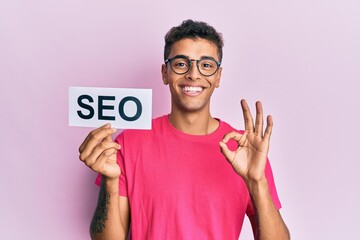 Young handsome african american man holding seo message paper doing ok sign with fingers, smiling...